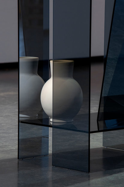 The showcase is emblematic of the presentation of the analog object in analog space: panes of glass that overlap and collide, forming a three-dimensional space within space - a stage through which the vase(c) is supported and protected. The dark tinted and one-sided mirrored panes, bring out the properties of glass. It is a play with transparency and reflection that influences the viewing moment. The shifting and twisting of individual panes of glass creates illusionistic moments and unexpected angles that can be discovered by walking around the showcase.